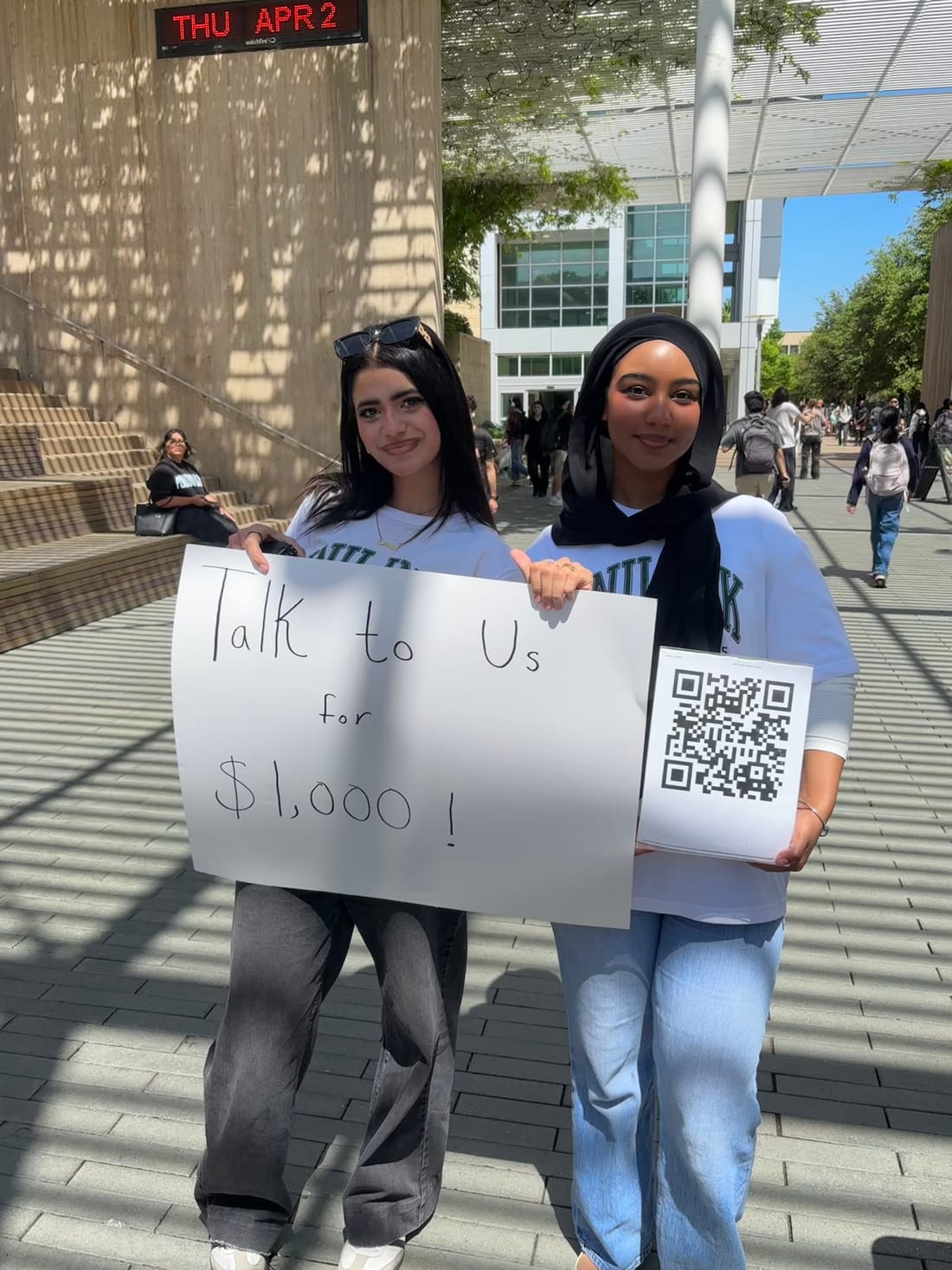 Two students on a campus plaza holding a hand-lettered 'Talk to Us for $1,000!' sign with a QR code.